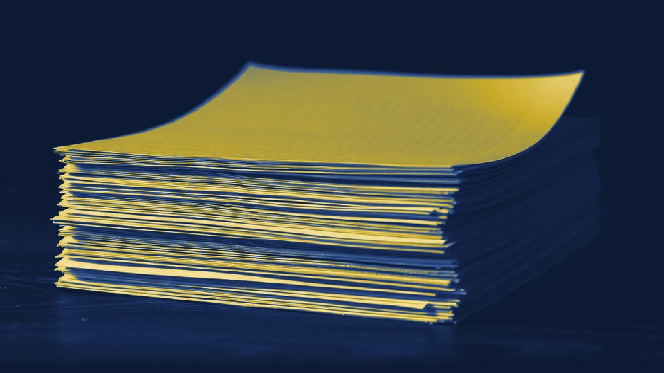 Uma pilha de doze folhas de papel pautado sobre uma mesa escura, com a folha de cima levemente deslocada